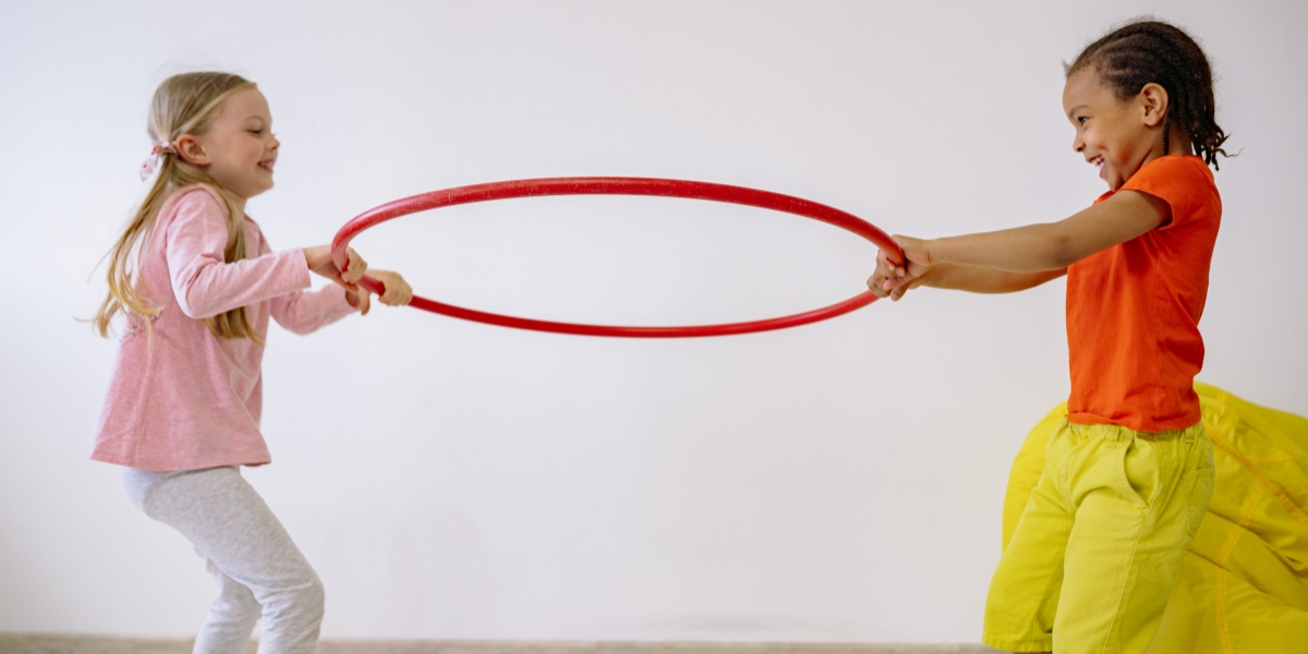 Two children hold a red hoop between them with their hands, smiling.