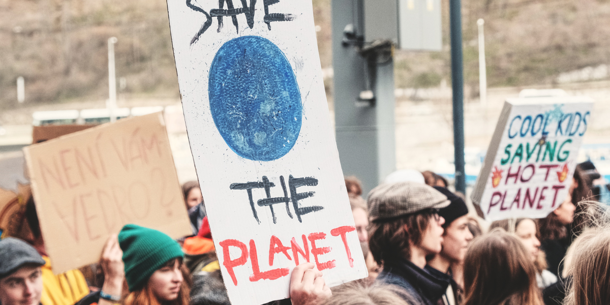 A group of people holding signs at a rally. One sign says "SAVE THE PLANET" with a blue drawing of the Earth. Another sign says "COOL KIDS SAVING HOT PLANET".