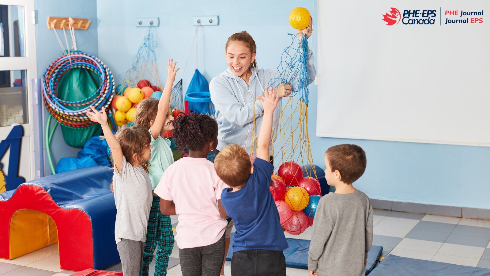 A teacher holding a net with balls in it in a room that seems to allow physical activity for pupils. She has 5 pupils in front of her, 3 girls and 2 boys. 3 of them raise their hands to get a ball from the teacher. / Une enseignante qui tient un filet avec des ballons dedans dans une salle qui semble permettre de l’activité physique à des élèves. Elle a en face d’elle 5 élèves, 3 filles et 2 garçons. 3 d’entres eux lèvent la main pour avoir une balle de la part de l’enseignante.