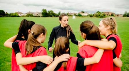 a group of 5 girls in a huddle facing their coach/teacher. They are wearing red pinnies and a black t-shirt while the adult is wearing a black track suit.