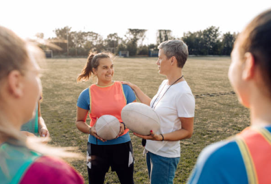 a teacher instructing their physical education class how to play rugby. the teacher is holding a rugby ball and the students are wearing orange pinnies. 
