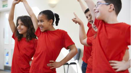 4 students standing in a dance studio wearing red shirts pointing to the ceiling and smiling