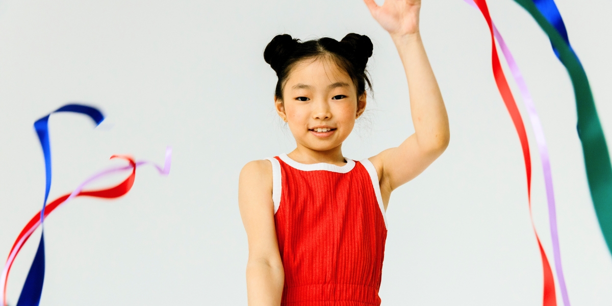 A young person in a red leotard smiles while holding up a hand and waving colorful ribbons against a white background. / Une jeune personne vêtue d'un justaucorps rouge sourit en levant la main et en faisant virevolter des rubans colorés sur fond blanc.