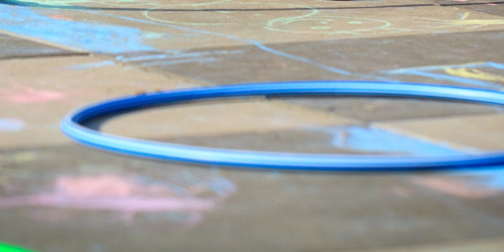 A child's legs wearing striped socks and red shoes are visible as they draw with chalk on a sidewalk covered in drawings and hula hoops.