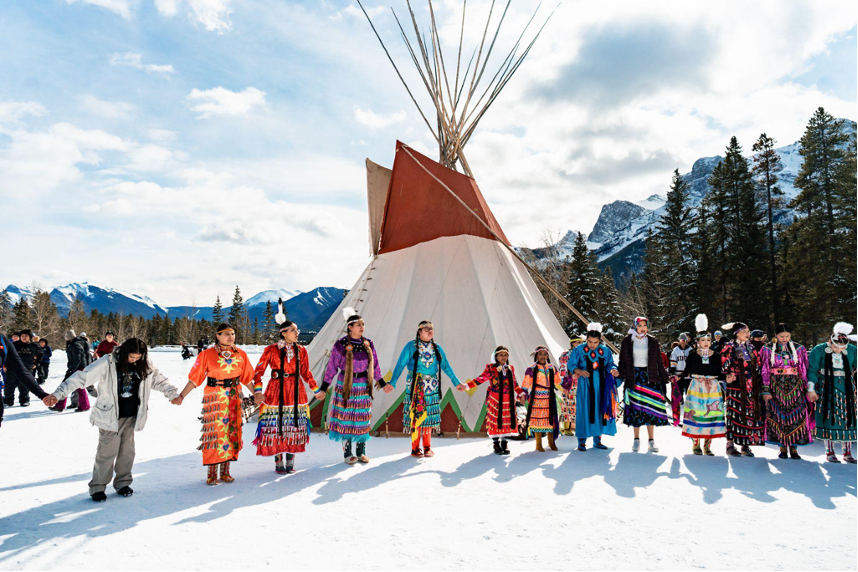 La photo montre un événement festif se déroulant sur un sol enneigé, célébré par des personnes vêtues d'habits traditionnels indigènes.