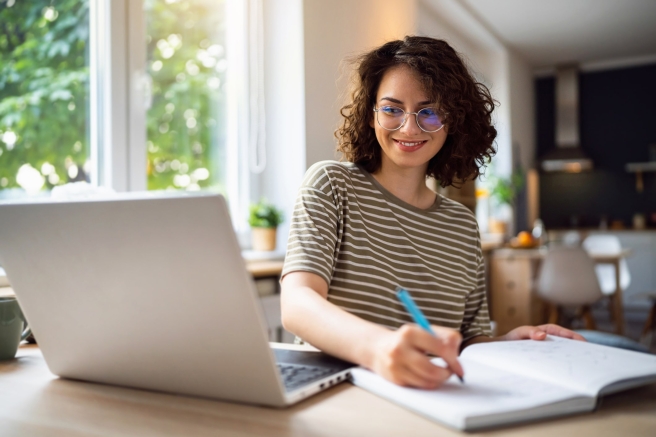 Une femme portant des lunettes est assise à une table, travaillant sur un ordinateur portable et un cahier à côté d'elle.