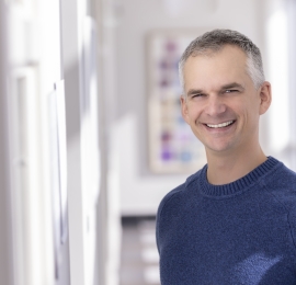 middle aged, white male, with buzz cut white and grey hair. He is wearing a navy blue crew neck knit sweater, and it standing in a hallway smiling.