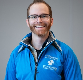 Homme blanc d'âge moyen, aux cheveux roux et à la barbe rousse, portant des lunettes noires et une veste imperméable bleu vif.