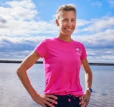 a middle-aged white woman, with blonde hair in a pony-tail, standing in a pink t-shirt with her hands on her hip infront of a lake