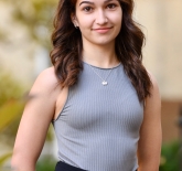 a woman with shoulder length brown hair with tanned skin tone. She is wearing a black adn white stripped tank top and dark blue pants. 