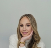 a white skin tone woman, with long blonde hair. She is wearing a white, long sleeved, cashmere shirt and a gold watch. She is sitting down, with her arms cross in fron of her with her chin resting in the pal, of her left hand.