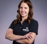 white women with shoulder length brown hair, smiling in the photo, she is standing with her arms crossed at her chest. She is wearing a black shirt and it has a small logo on the left chest that says "Beyond the Win - Education Program"