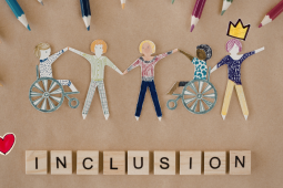 Paper cutouts of diverse people, including some in wheelchairs, holding hands, surrounded by colored pencils. Wooden letter blocks spell out 'INCLUSION'.
