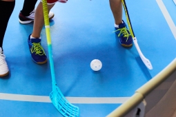 two indoor hockey sticks on the ground in a gymnasium which has blue flooring. There is a child holding each stick - you can only see their shoes. They are trying to hit a white whiffle ball. 