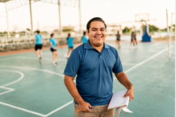 a physical education teacher standing on an outdoor basketball court holding a clipboard. They are wearing a blue polo, grey pants and are smiling at the camera. In the background you can see students playing basketball. 