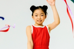 A young person in a red leotard smiles while holding up a hand and waving colorful ribbons against a white background. / Une jeune personne vêtue d'un justaucorps rouge sourit en levant la main et en faisant virevolter des rubans colorés sur fond blanc.