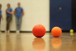 two red dodgeballs on the floor of a gymnasium with 3 students standing against a wall