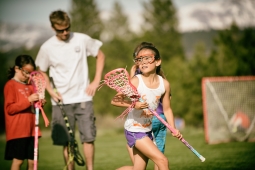 two children playing lacrosse. They are wearing eye goggles and holding a lacrosse stick. There is an adult and a net in the background. 