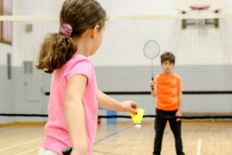 a young girl and boy playing badminton. The girl has the shuttle cock and is wearing a pink shirt with her hair in a pony tail. The boy is standing on the other side of the net wearing an orange shirt, black pants, and is waiting for the birdie with his racket up. 