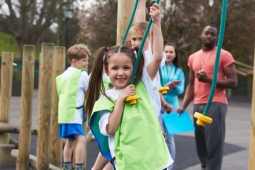 4 young students playing outside on a rope with a teacher in the background. The children are wearing a green pinnie and the teacher is wearing a red t-shirt. 