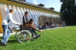 a child in a wheelchair throwing a ball with another peer behind them holding the handles of their chair. There is a teacher beside them and they are on a grass field.