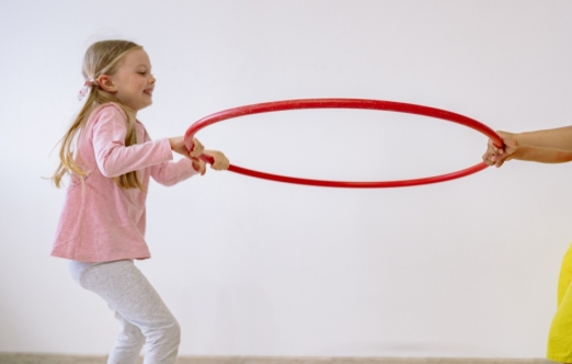 Two children hold a red hoop between them with their hands, smiling.