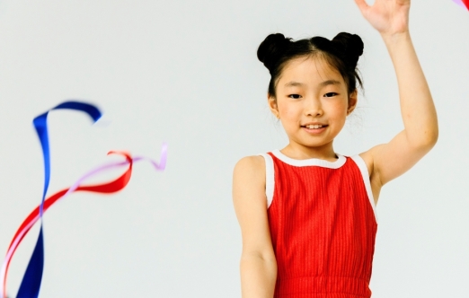 A young person in a red leotard smiles while holding up a hand and waving colorful ribbons against a white background. / Une jeune personne vêtue d'un justaucorps rouge sourit en levant la main et en faisant virevolter des rubans colorés sur fond blanc.