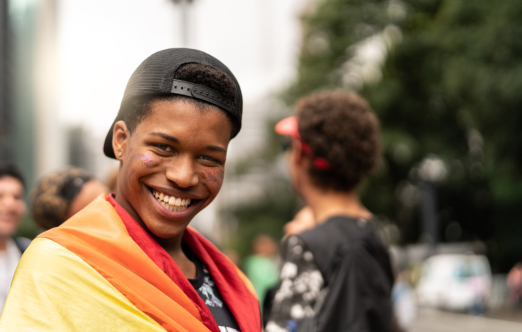 Une jeune personne portant un drapeau arc-en-ciel et affichant un sourire confiant et contagieux.
